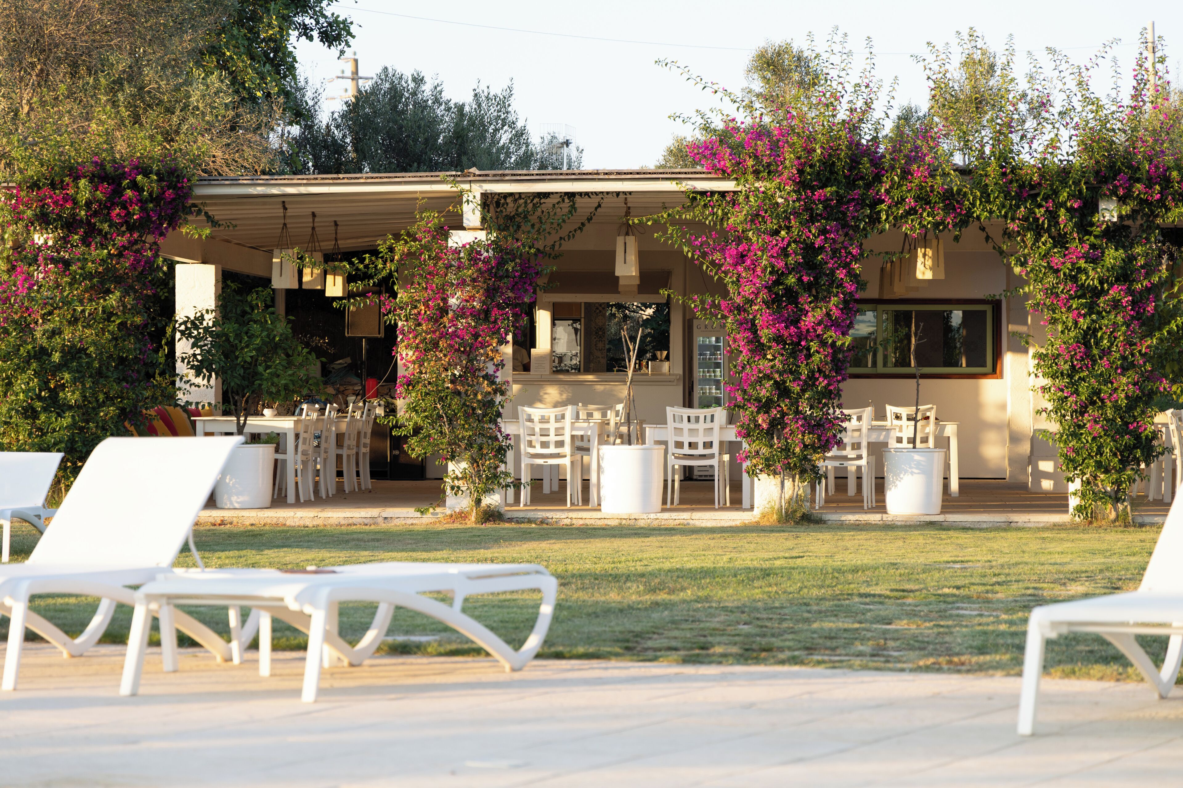 Un patio al aire libre con sillones blancos, un espacio de estar cubierto y plantas con flores.