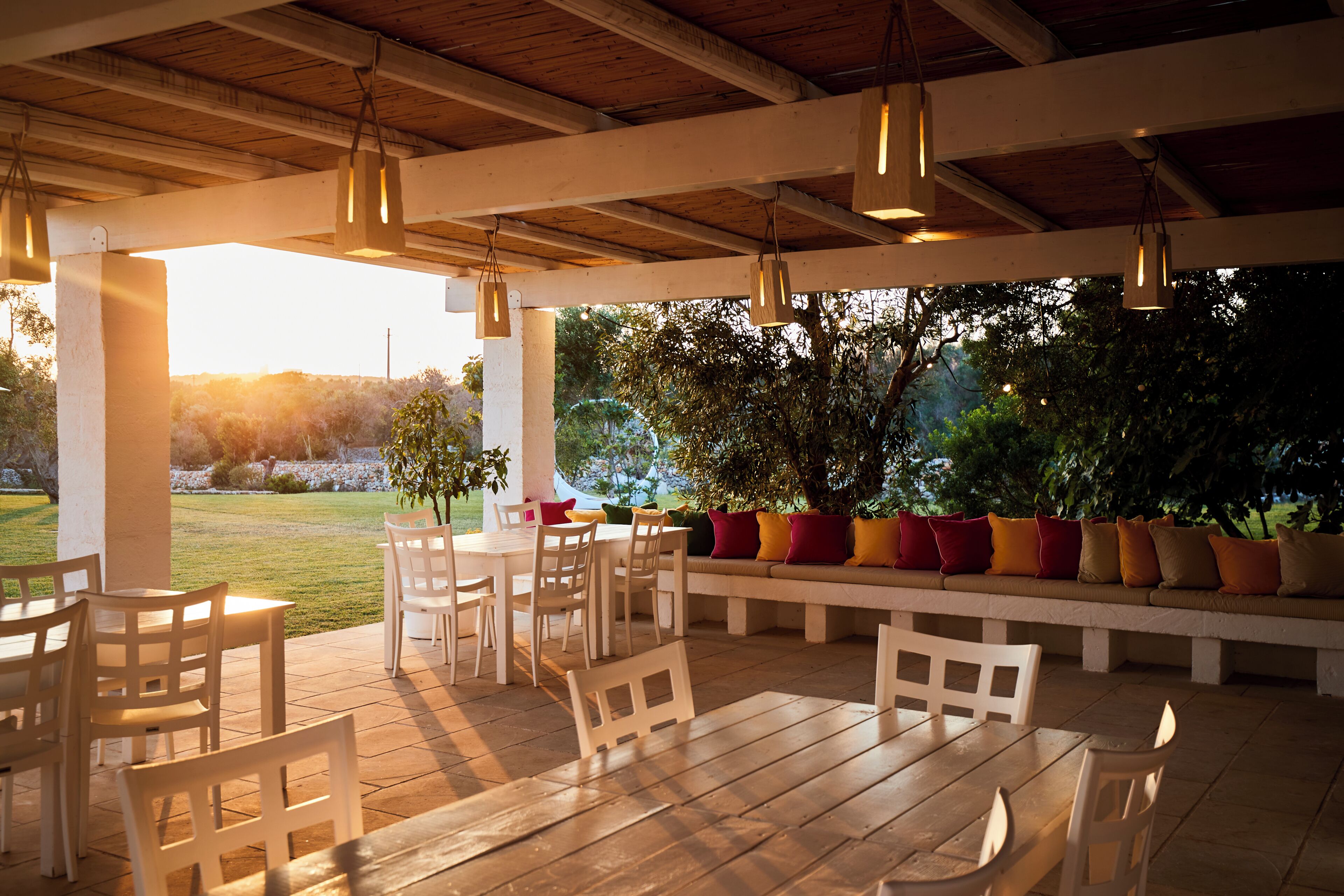 An outdoor patio area with wooden tables and chairs, a bench with colorful cushions, and hanging pendant lights.
