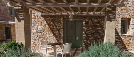Un edificio de piedra con una pérgola de madera, mobiliario exterior y una puerta con contraventanas verdes.