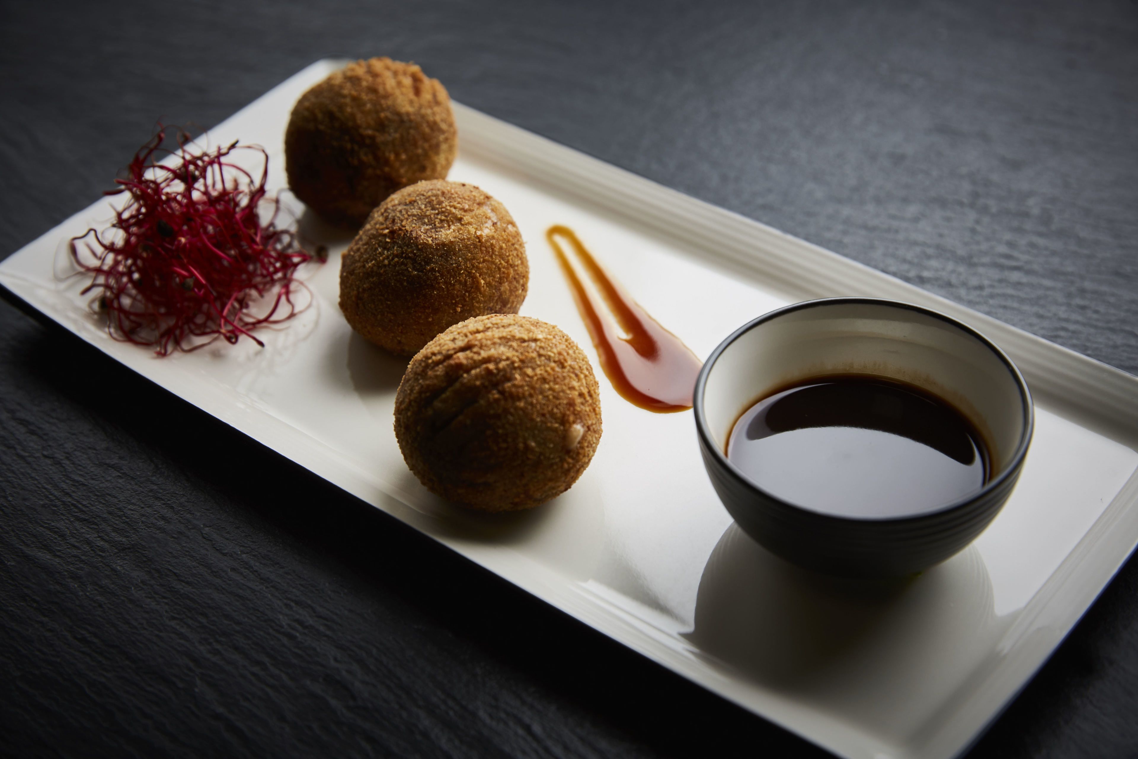 A plate with three round, breaded items, a small bowl of sauce, and a pile of red garnish on a dark surface.