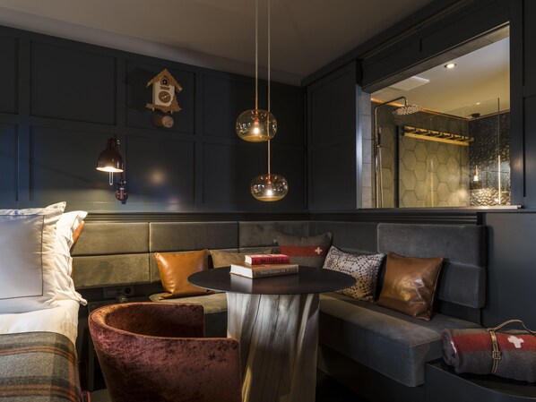 A modern hotel room with a dark color scheme, a cozy seating area, and a large mirror reflecting the bathroom. - HUUS Gstaad (Saanen)