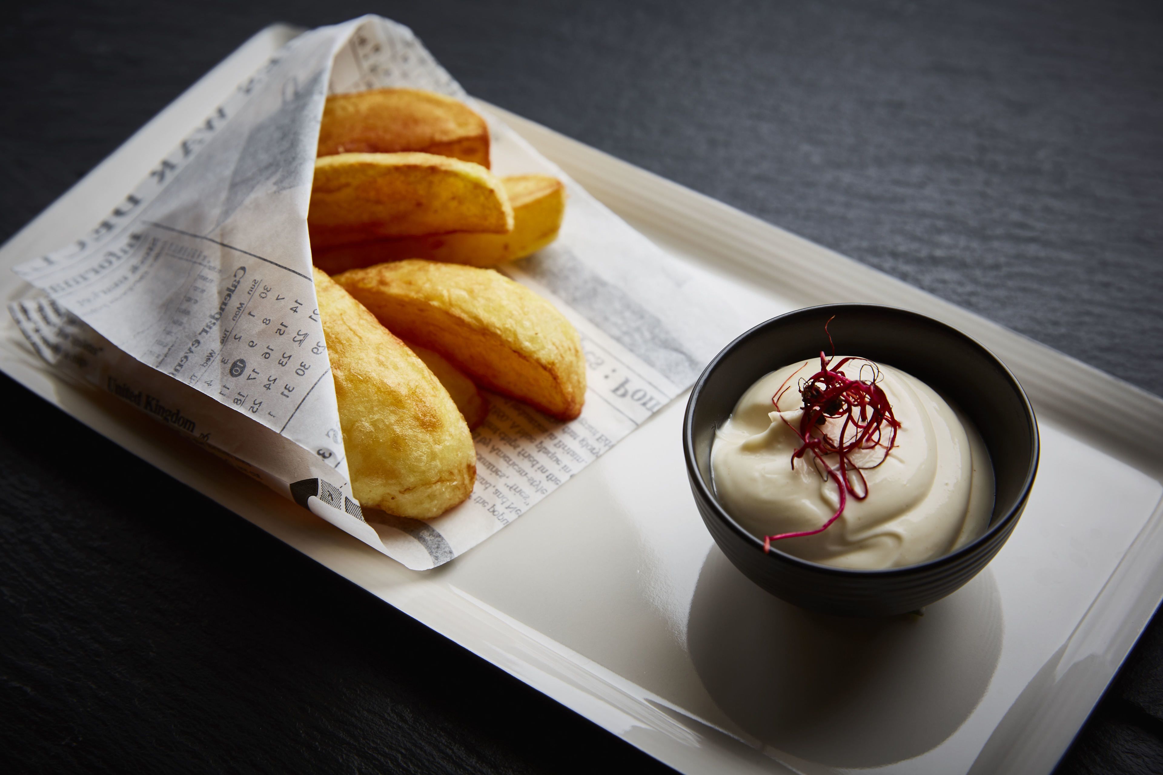 A tray with golden-brown fried items and a bowl of white dip with red garnish on a dark surface.