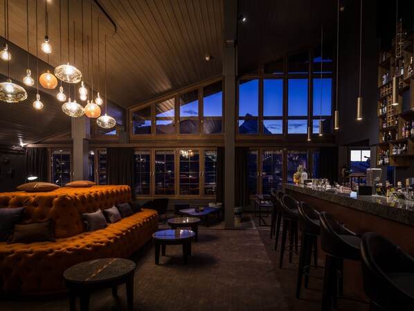A spacious bar area with a high ceiling, large windows, and a variety of seating options including a sofa and round tables. - HUUS Gstaad (Saanen)