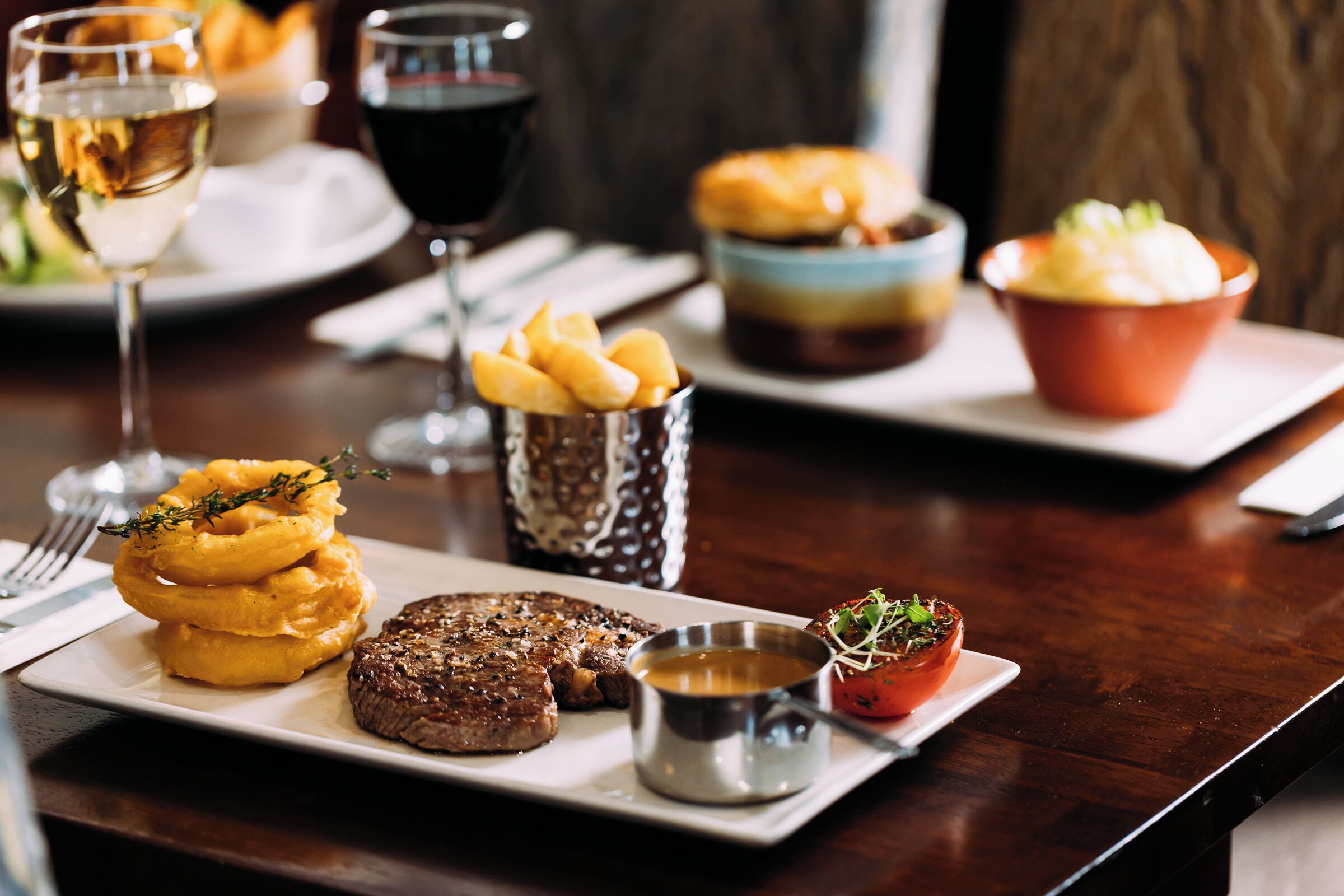 A dining table with a steak, onion rings, and a small bowl of sauce.