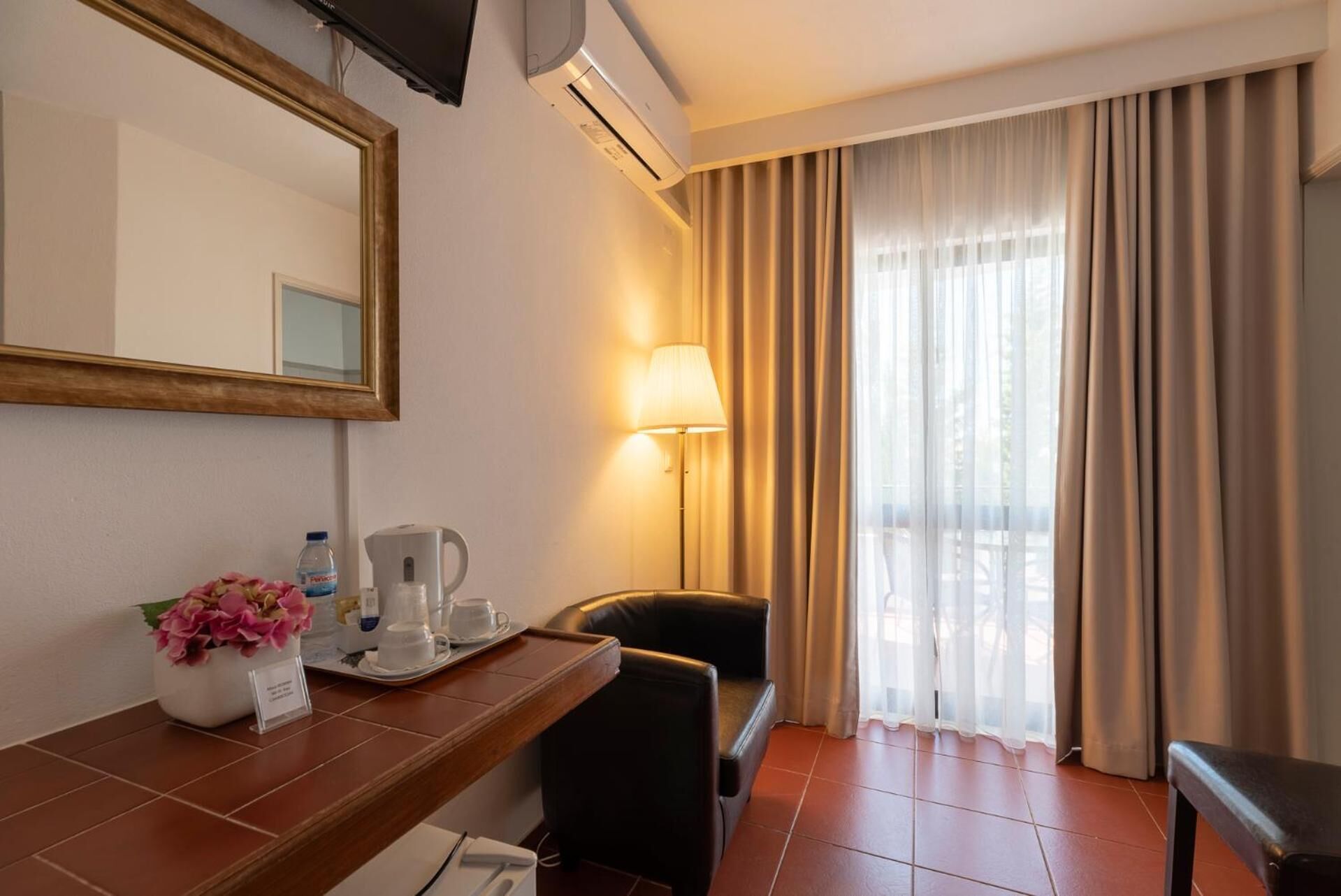 Hotelkamer met een houten bureau, een stoel, nachtkastje met lamp, spiegel en raam met gordijnen.