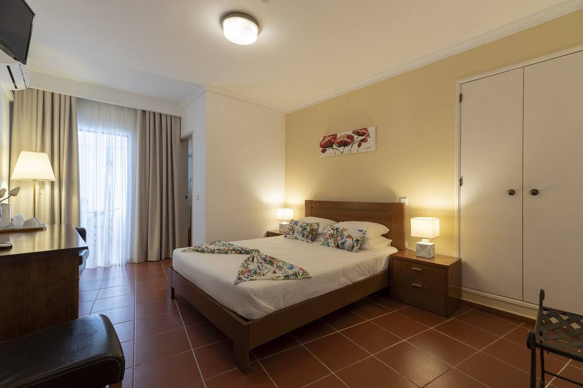 A hotel room with a bed, bedside tables, a desk, a chair, a wardrobe, and a window with curtains.