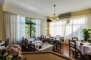 Uma sala de jantar com mesas preparadas para o serviço, grandes janelas com cortinas e um ventilador de teto.