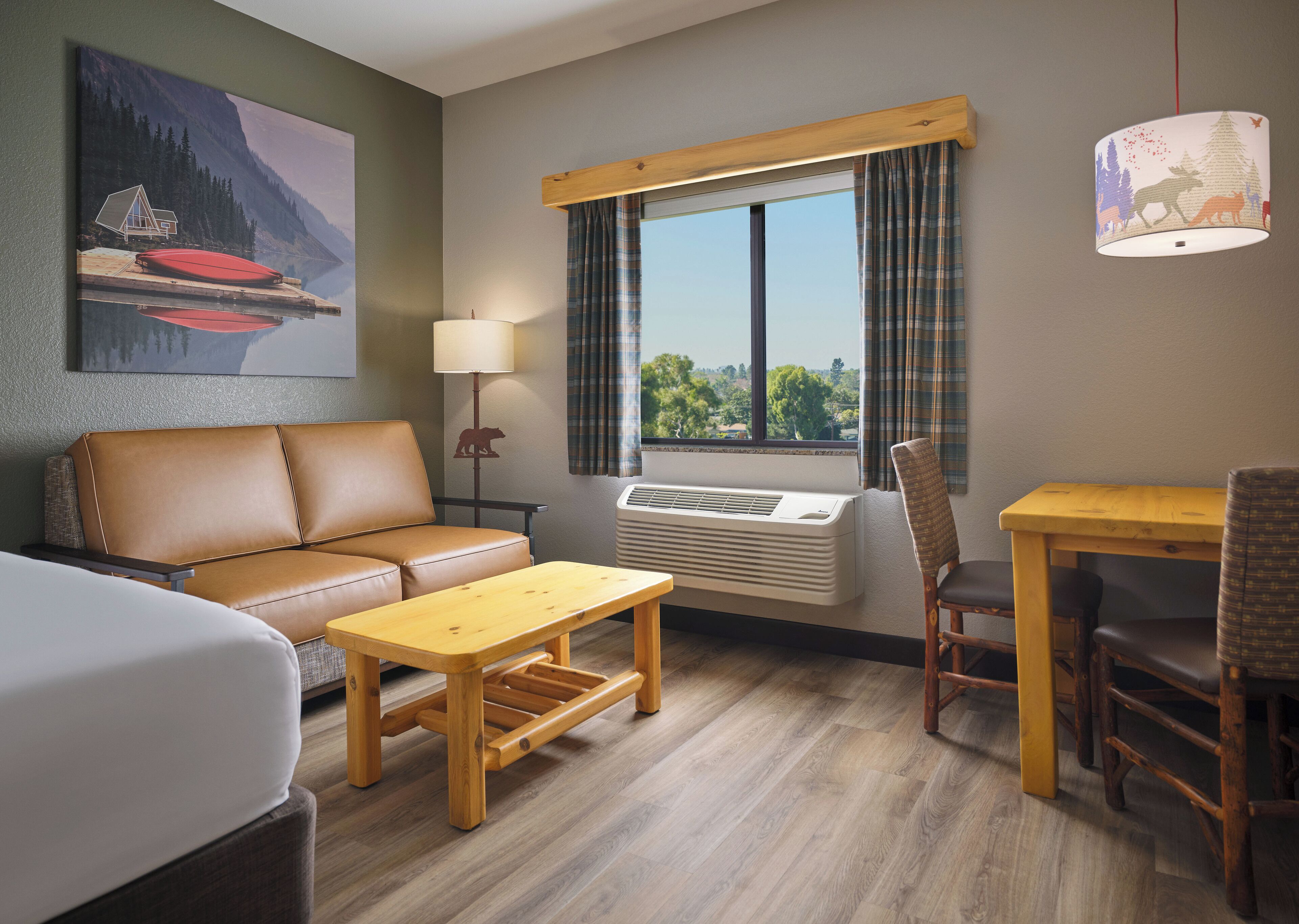 A hotel room with a sofa, a small table, a chair, a lamp, an air conditioning unit, and a window with curtains.