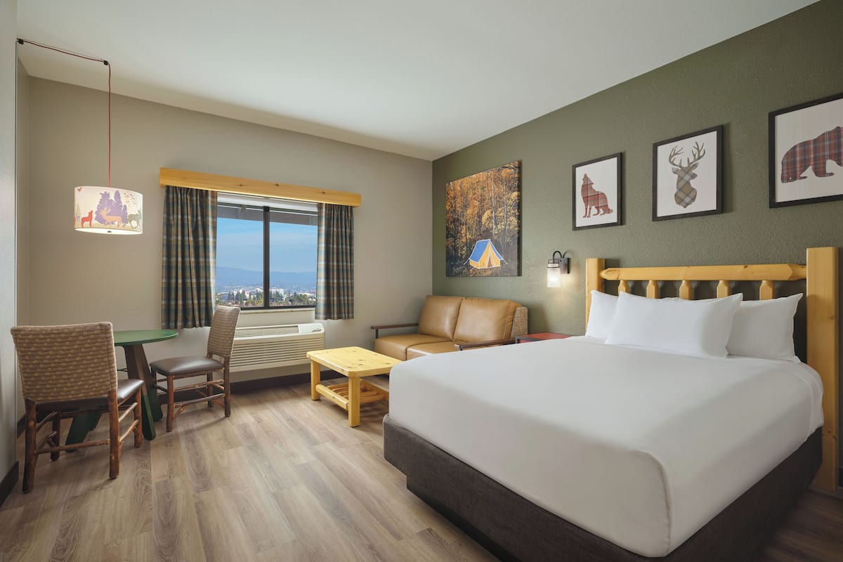 A hotel room with a large bed, a desk with two chairs, a sofa, and a window with a view. There are framed pictures on the wall and a lamp on the desk.