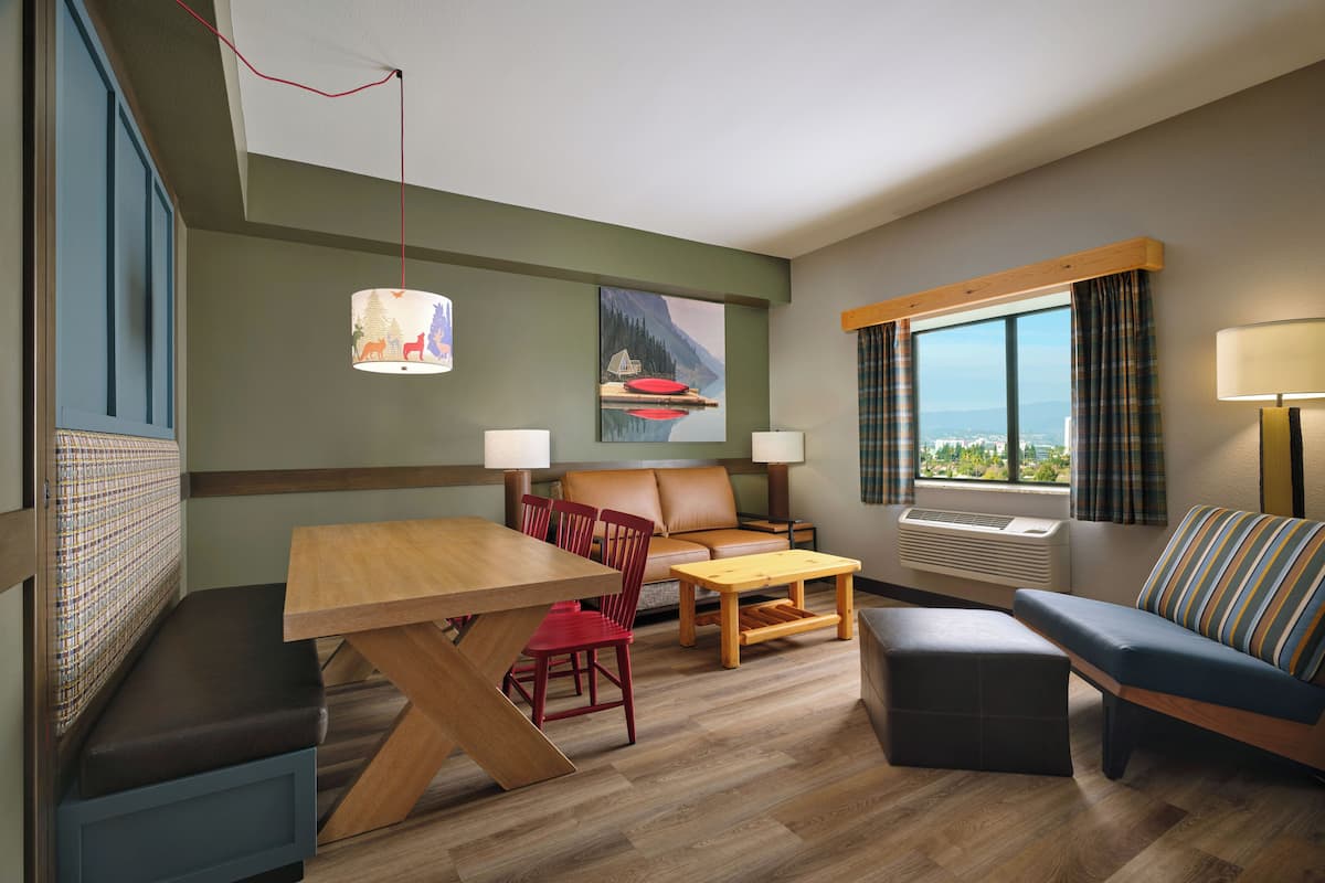 A hotel room with a dining table, red chairs, a sofa, a window with curtains, and a painting on the wall.