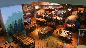An indoor restaurant with multiple wooden tables and chairs, a kitchen area, and a mural of trees on the wall. - Great Wolf Lodge Anaheim, CA (Garden Grove)