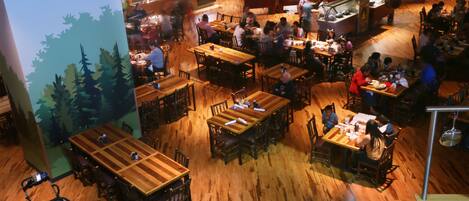 An indoor restaurant with multiple wooden tables and chairs, a kitchen area, and a mural of trees on the wall.