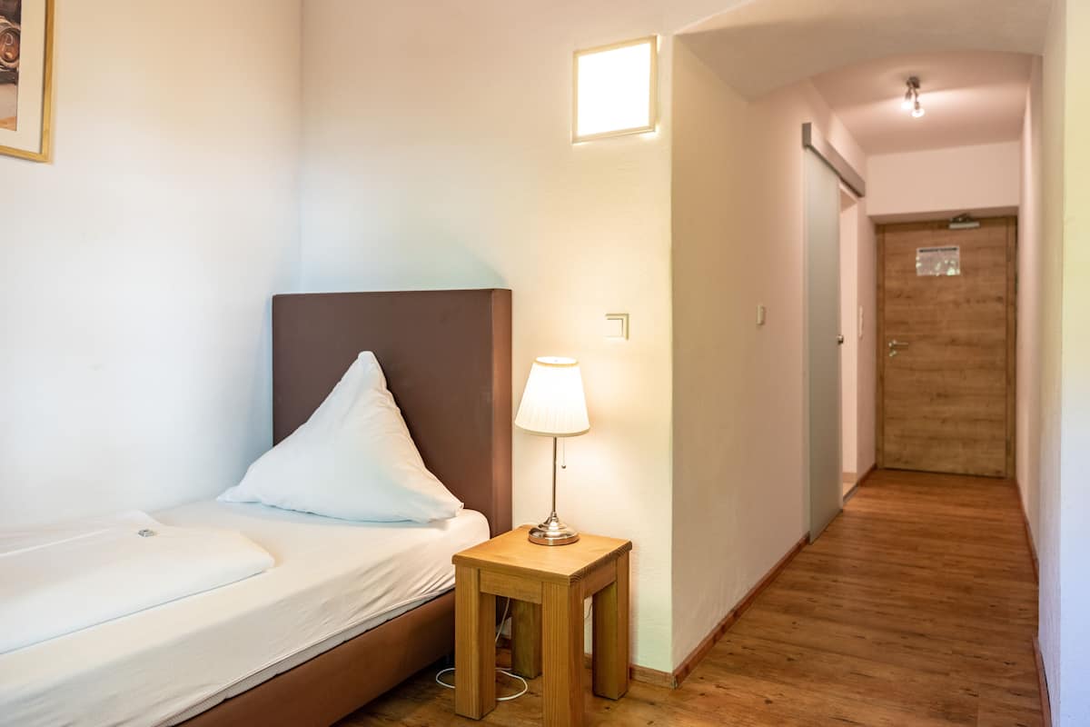 A hotel room with a bed, a bedside table with a lamp, a wooden floor, and a door leading to another room.
