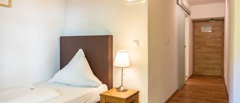 A hotel room with a bed, a bedside table with a lamp, a wooden floor, and a door leading to another room.
