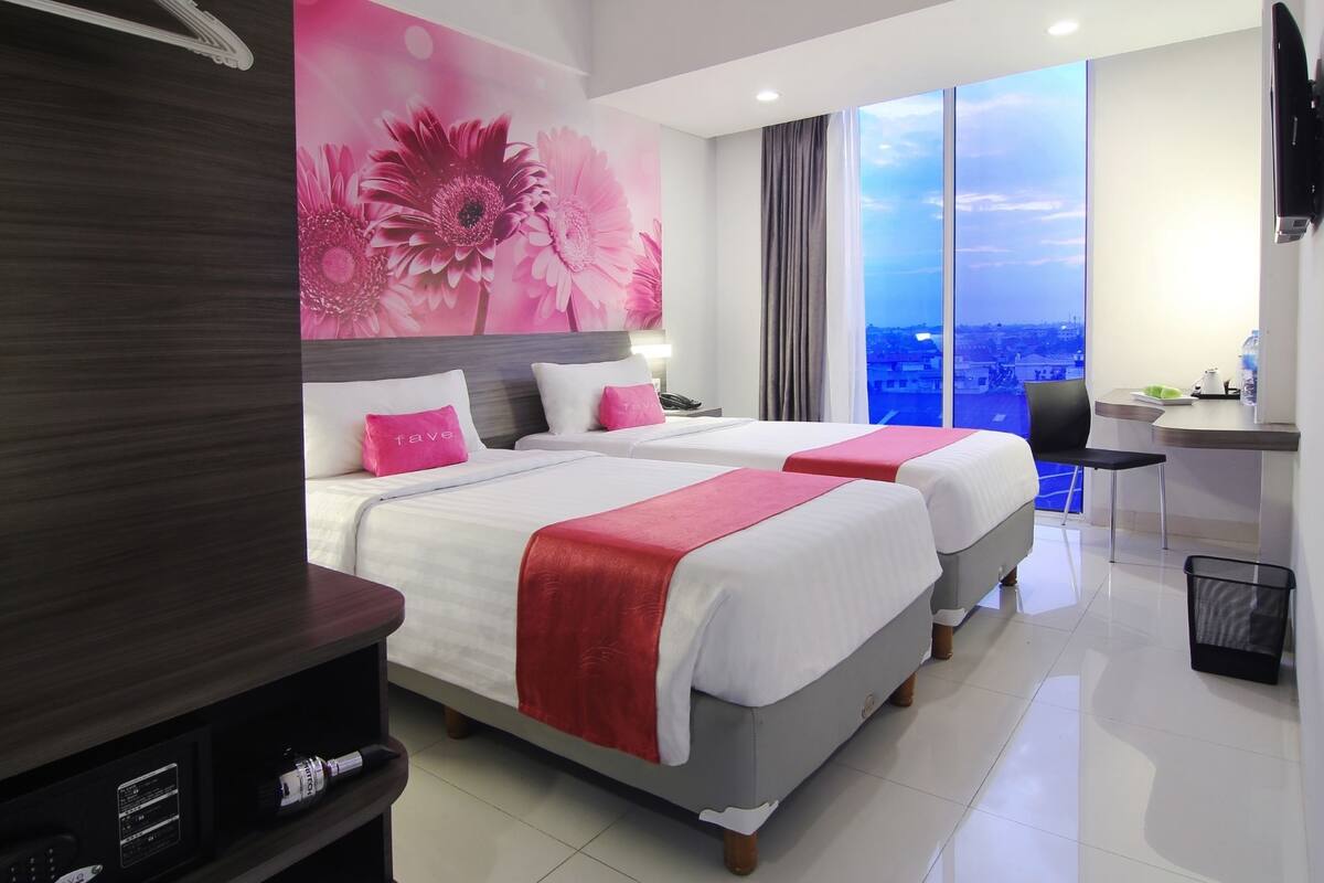 A hotel room with two beds, a desk, a chair, and a large window with a city view.