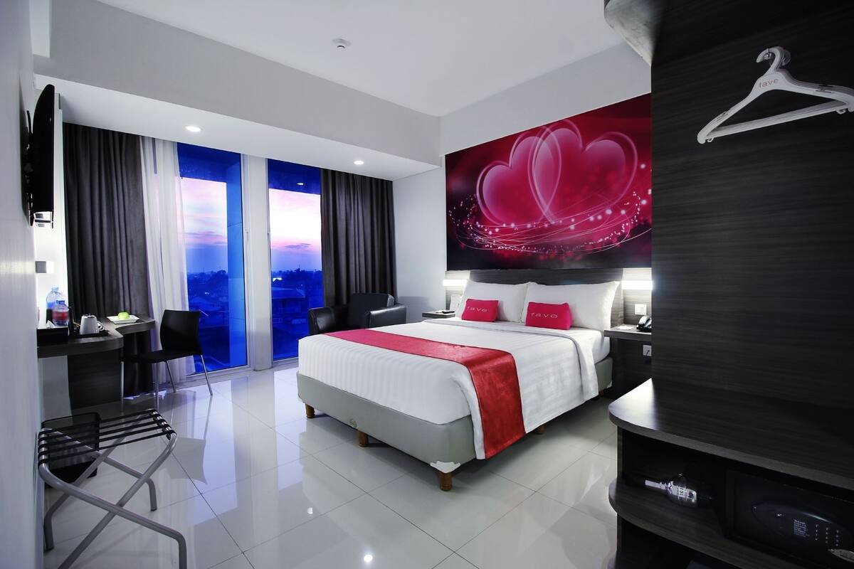 A modern hotel room with a large bed, a desk with a chair, a TV, and a view of the cityscape through the windows.