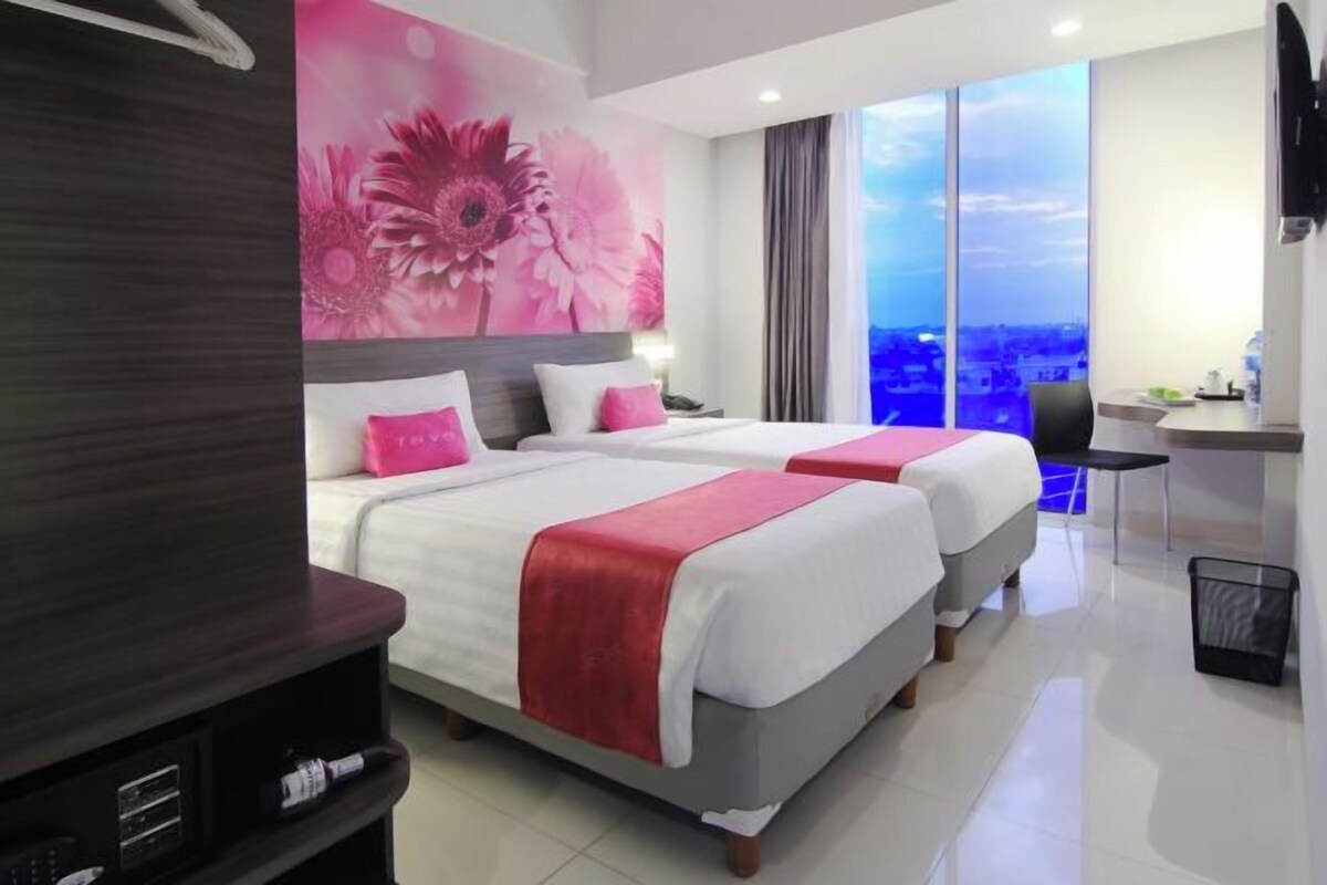 A hotel room with two beds, a desk, and a large window offering a city view.