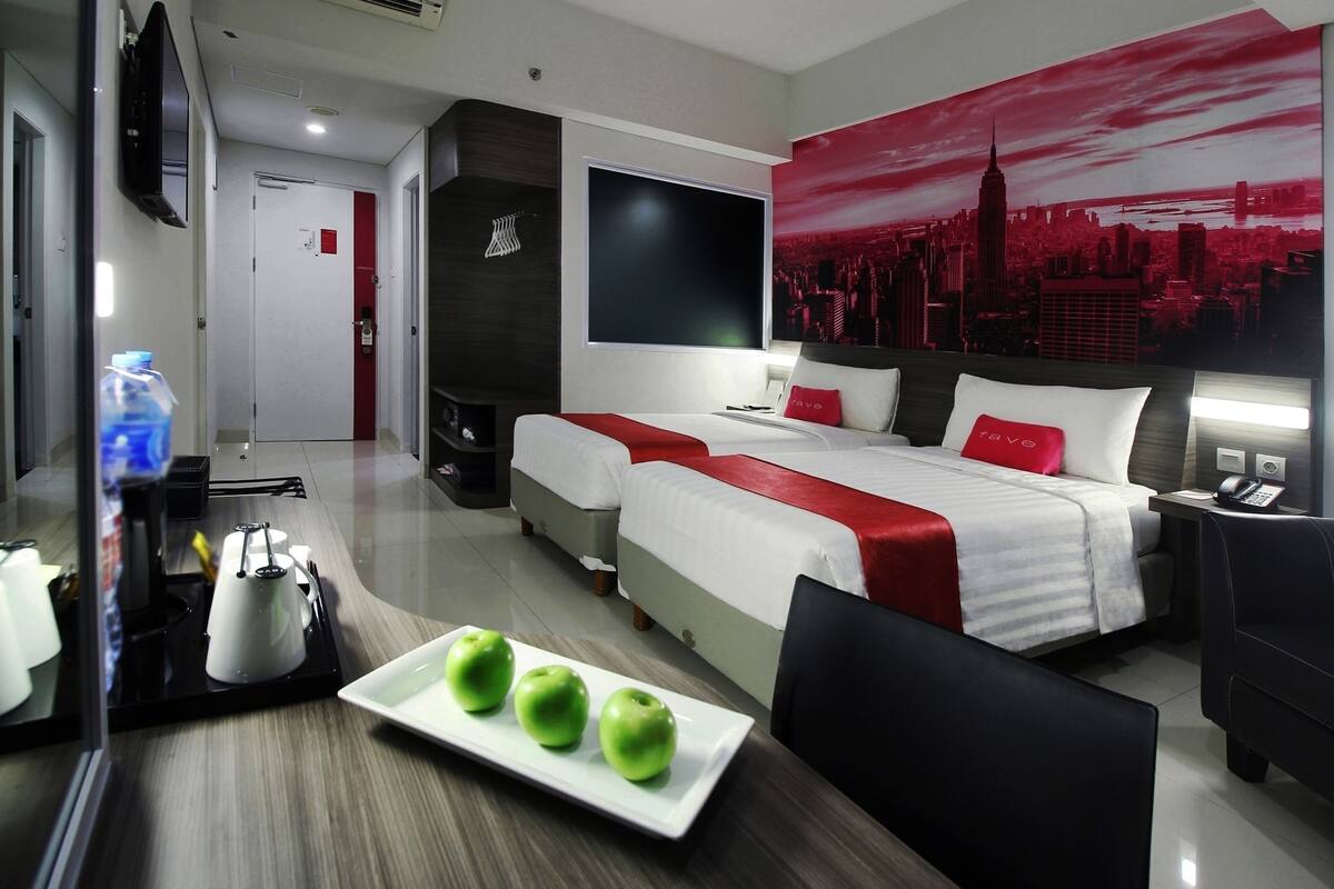 A hotel room with a bed, bedside tables, a TV, a red and white bedspread, and a cityscape mural.