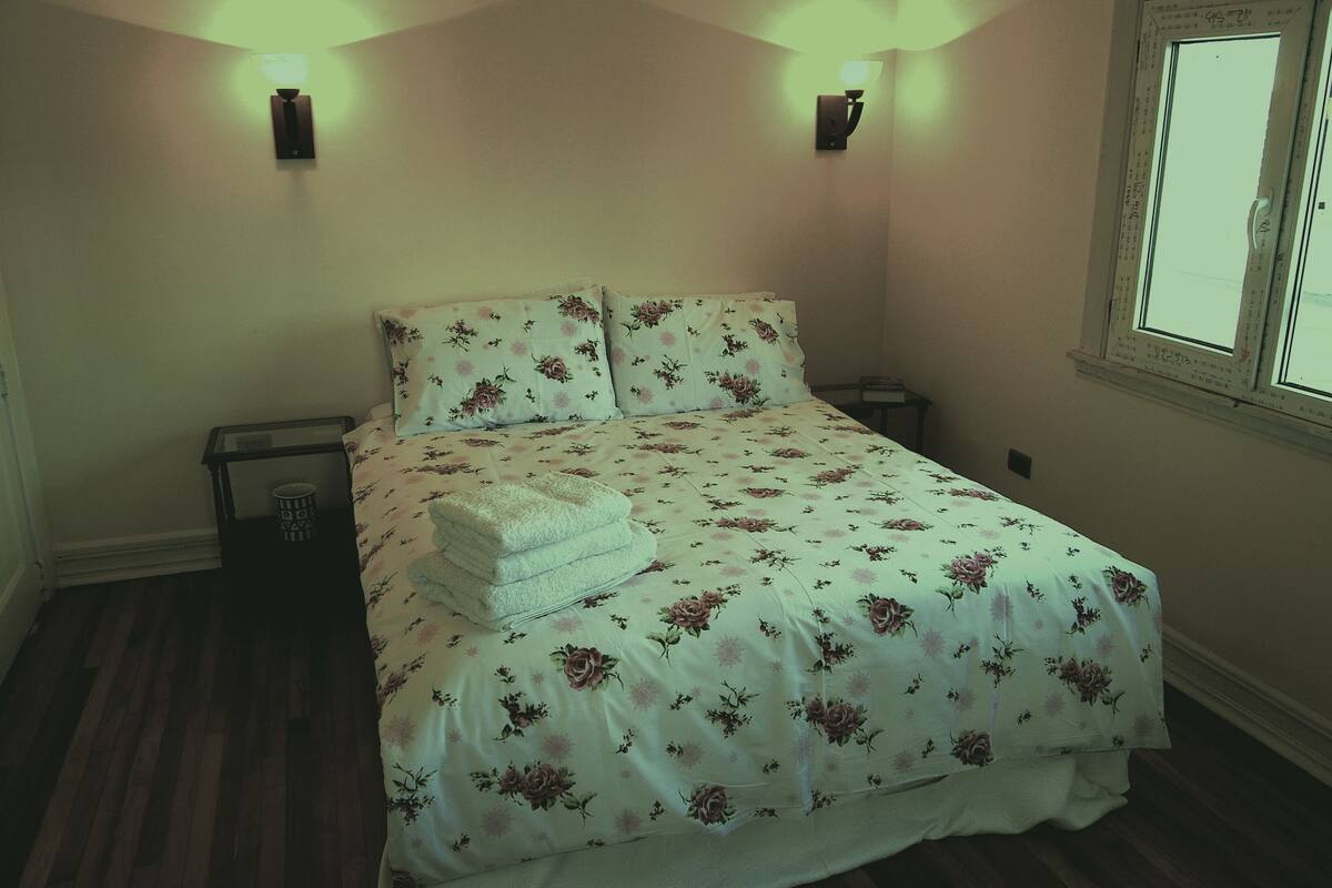 Quarto com edredom floral, duas luminárias de cabeceira, janela com vista e mesa de cabeceira com abajur.