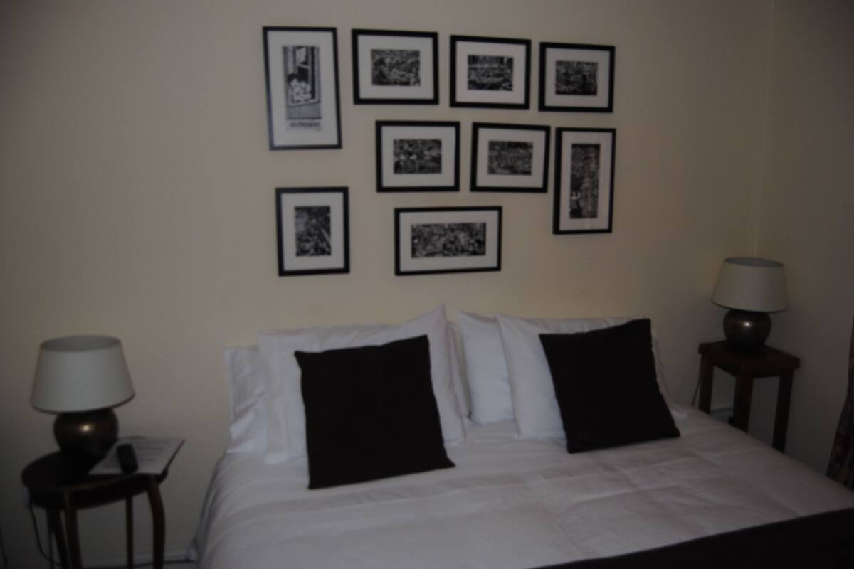Quarto de hotel com uma cama, duas mesas de cabeceira com luminárias e nove quadros na parede.