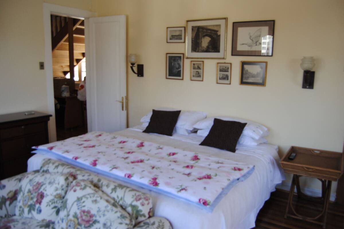 Quarto com cama, mesa de cabeceira, parede com quadros e porta que dá para outro cômodo.