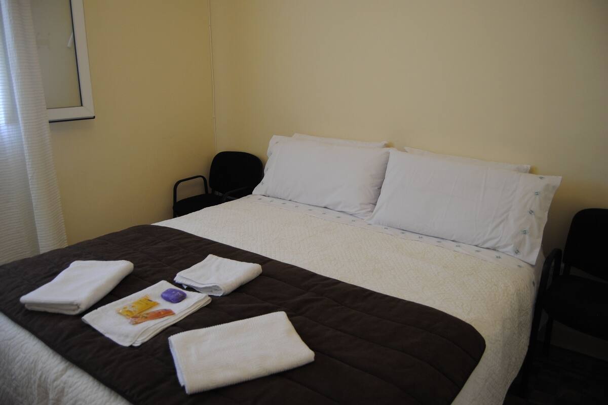 Cama bem arrumada com roupa de cama branca e castanha, uma cadeira preta e uma pequena mesa com artigos de higiene pessoal.