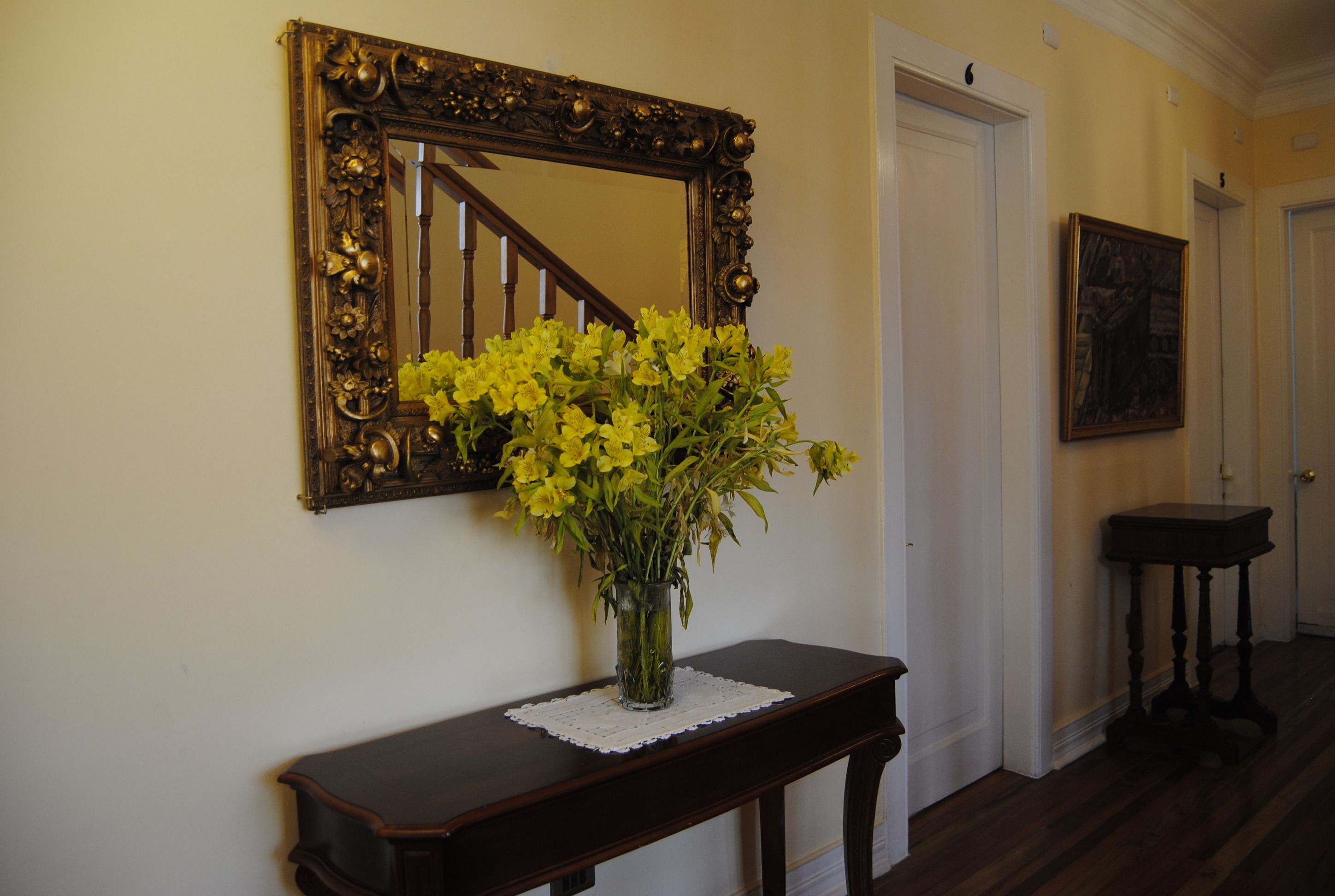 Un couloir avec une console en bois, un vase rempli de fleurs jaunes, un miroir doté d’un cadre orné et un tableau accroché au mur.