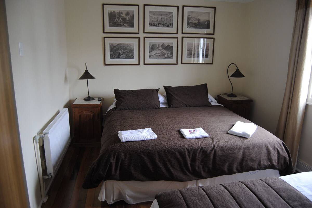 Cama bem arrumada com colcha marrom, duas mesas de cabeceira com abajures e seis quadros na parede.
