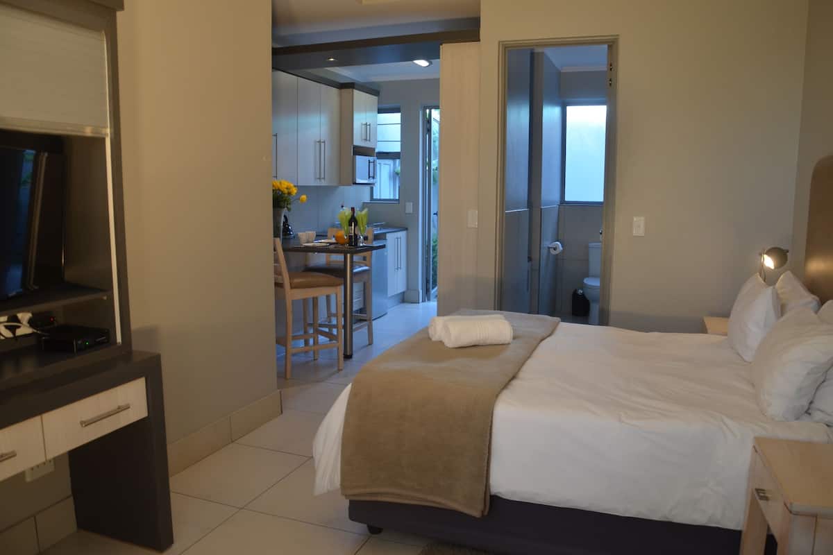 Ein Hotelzimmer mit Bett, Fernseher, Schreibtisch mit Stuhl und einer sichtbaren Küchenzeile durch eine offene Tür.