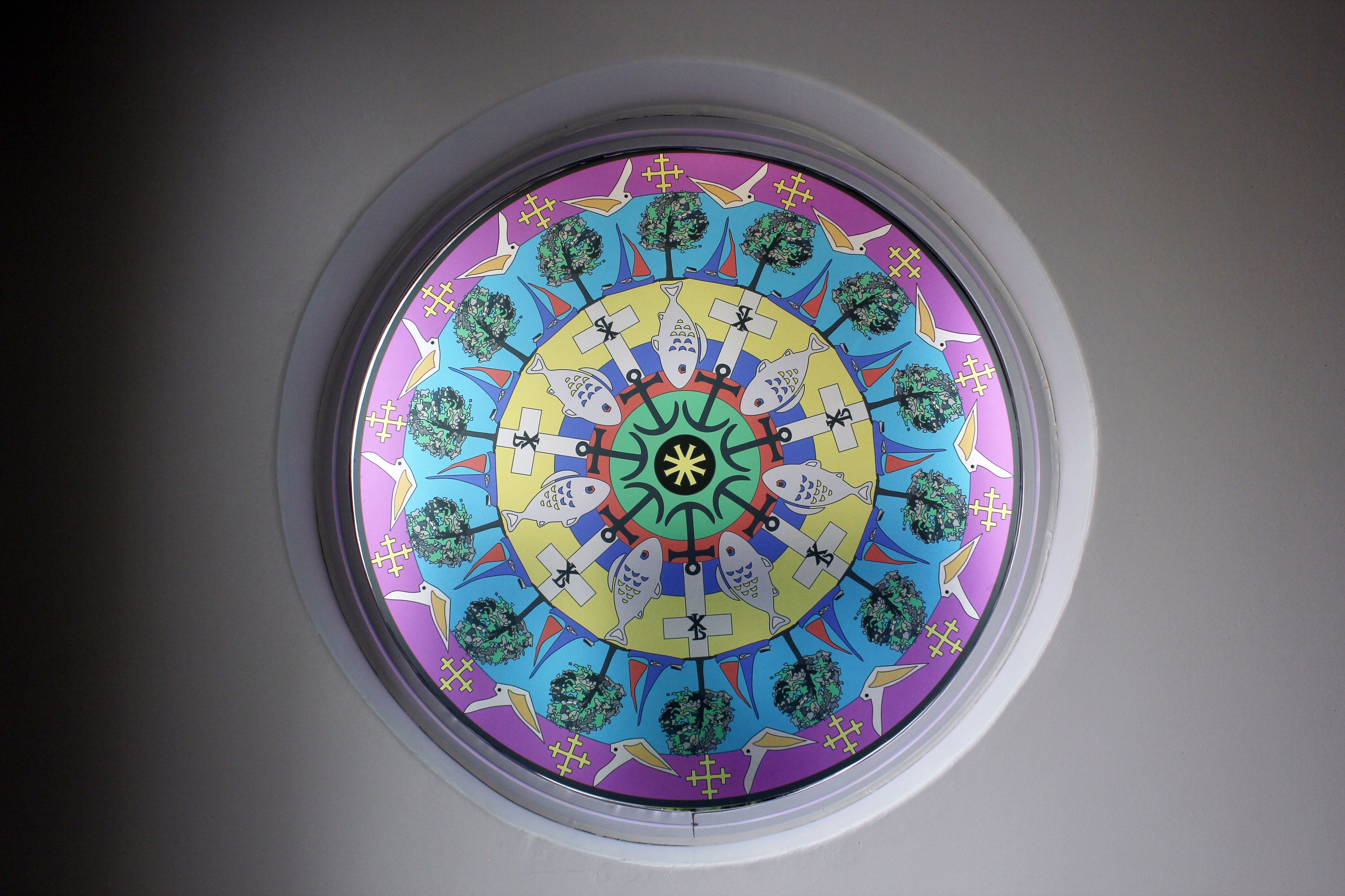 A circular stained glass window with a symmetrical design featuring fish, trees, and geometric patterns.