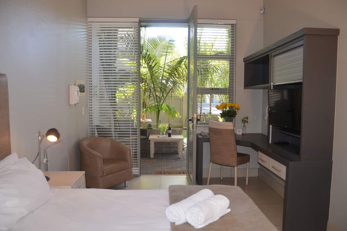 Ein Hotelzimmer mit einem Bett, zwei Stühlen, einem Schreibtisch mit Fernseher und Blick auf Palmen durch die Tür.