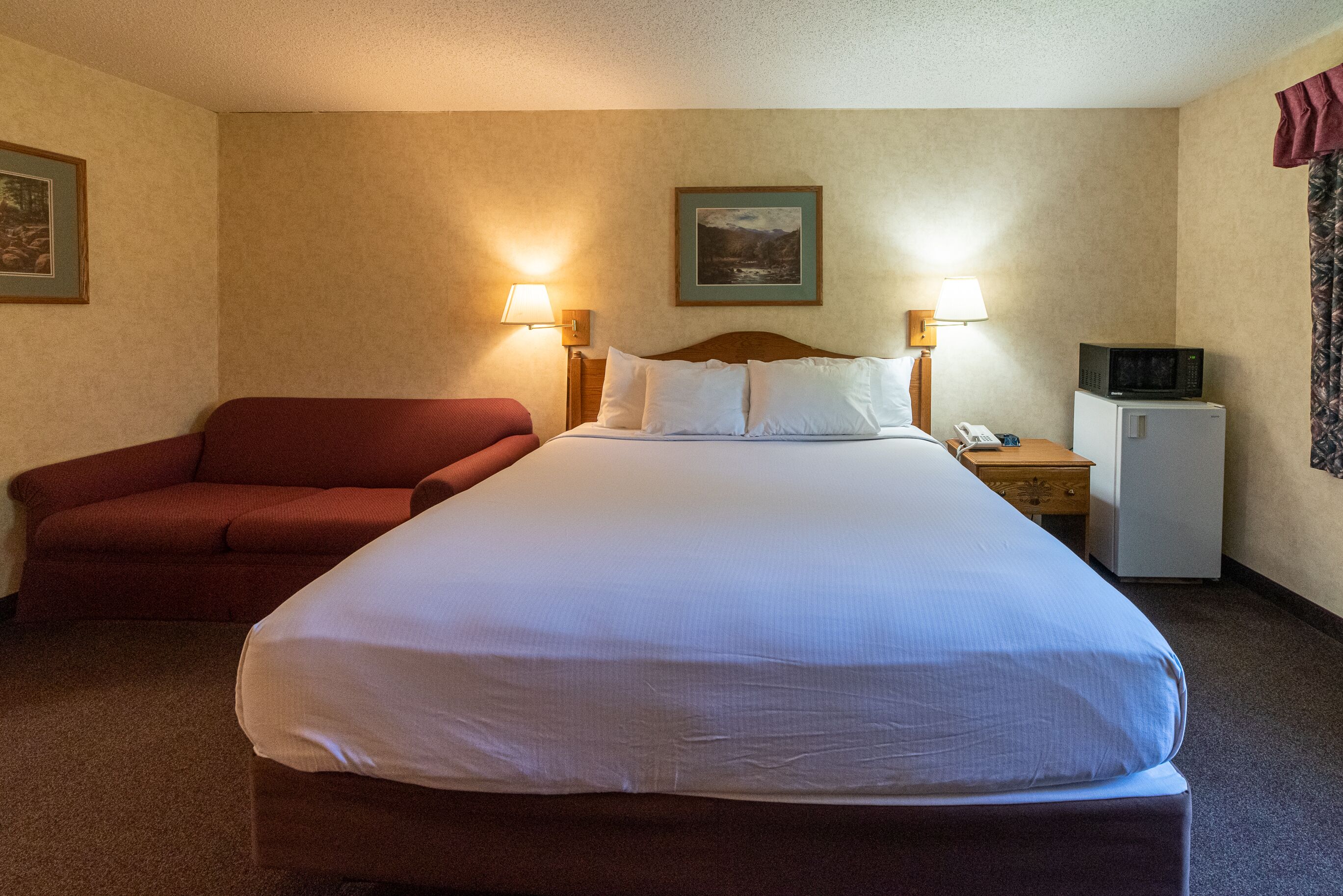 Camera d'albergo con un letto grande, un divano rosso, un piccolo frigorifero e due lampade a parete.