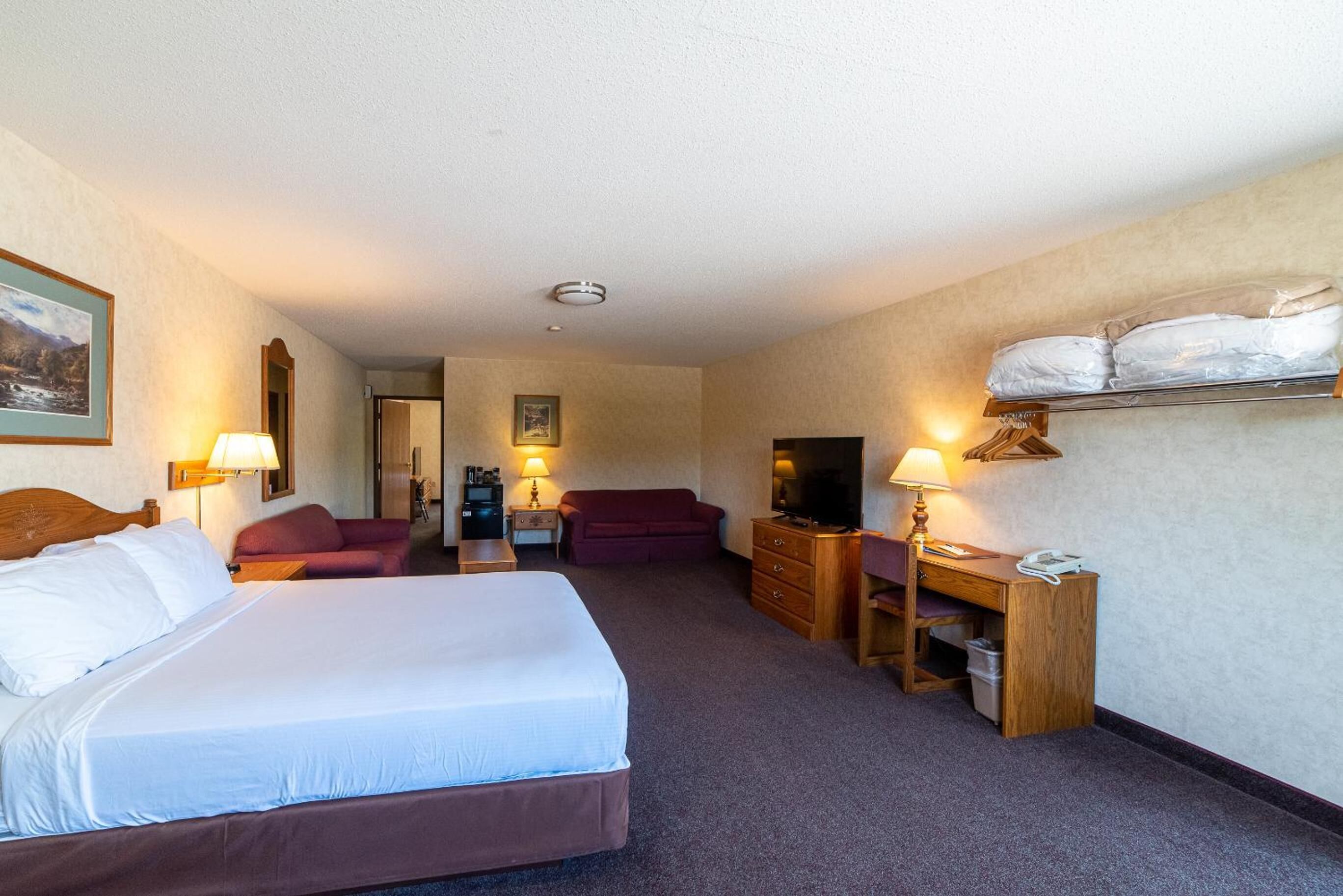 Une chambre d’hôtel avec un grand lit, un bureau avec une chaise, un canapé, une télévision et une étagère avec des coussins.