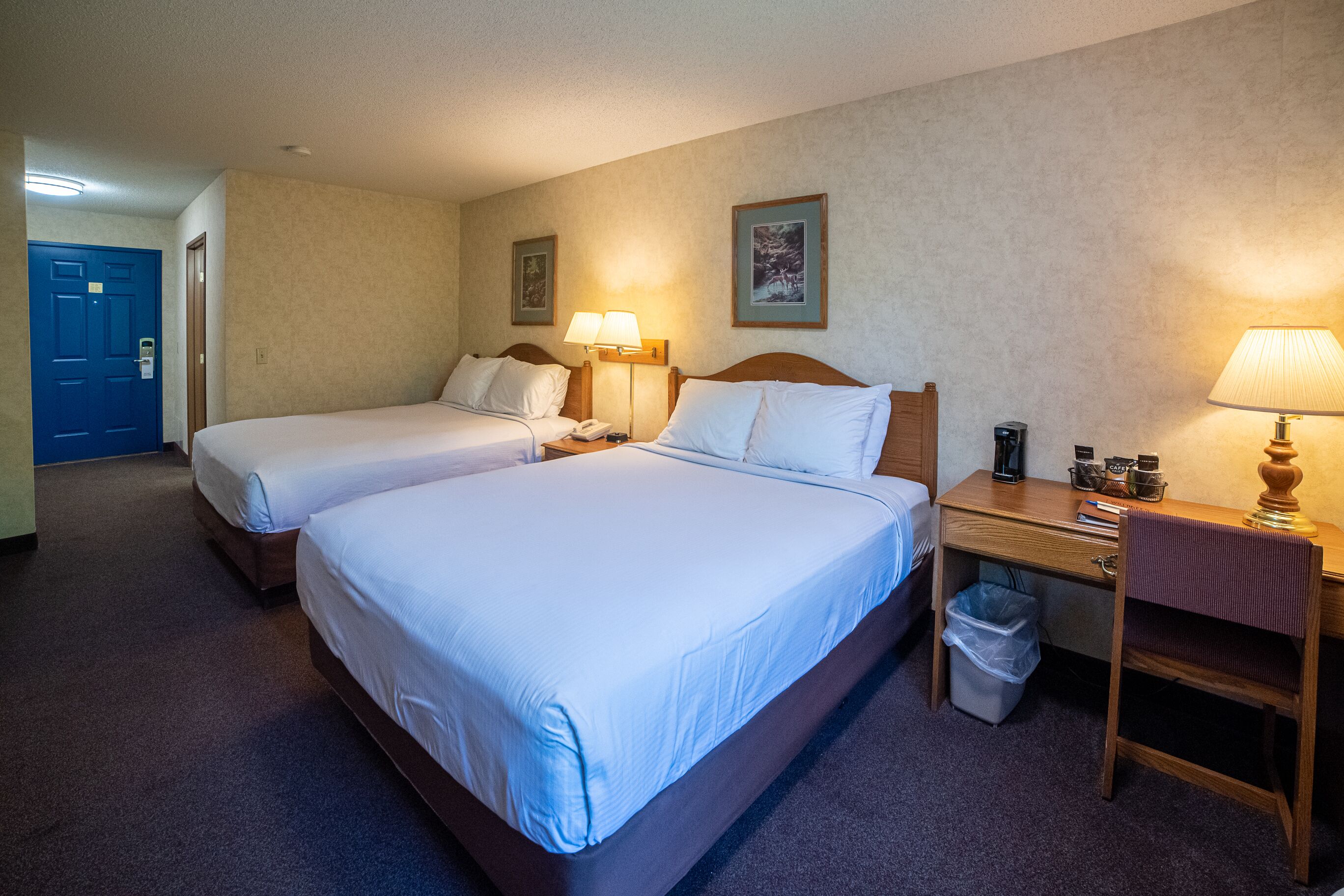 Quarto de hotel com duas camas, uma mesa com luminária, um cesto de lixo e uma porta.