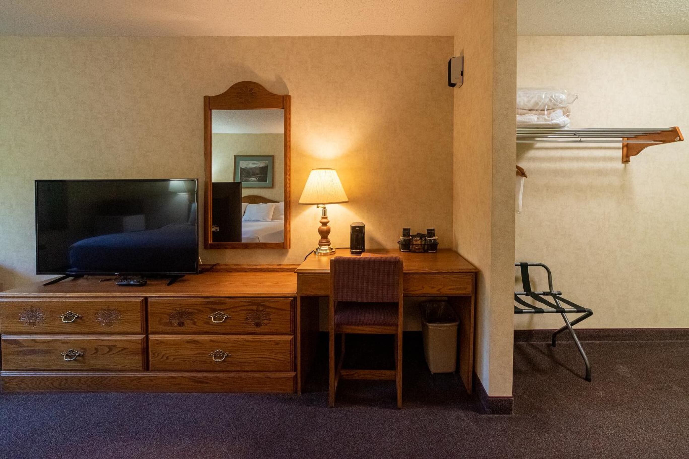 A hotel room with a wooden desk, a television, a lamp, a chair, a mirror, and a small shelf with toiletries.