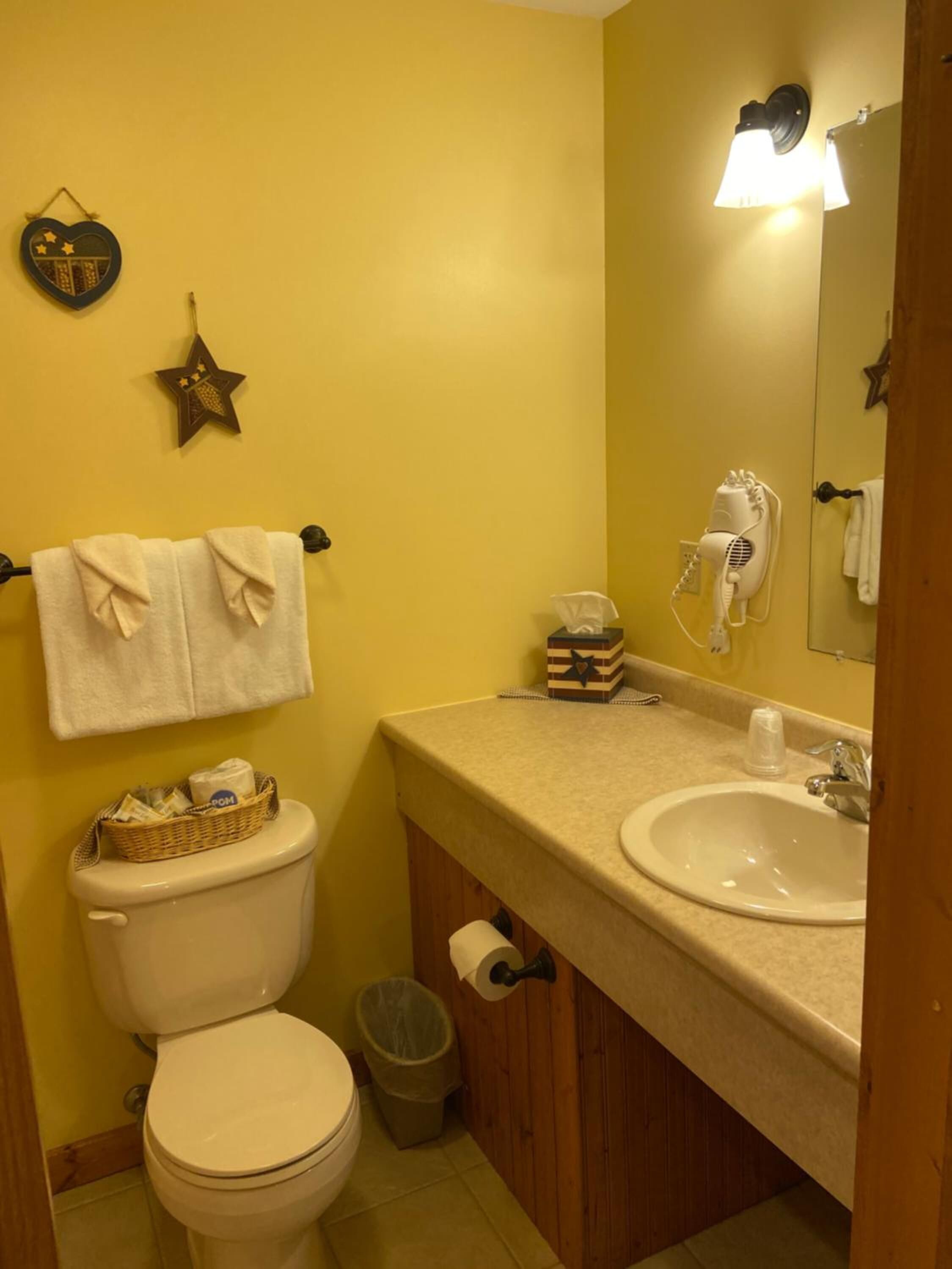 Une petite salle de bain comprenant des toilettes, un lavabo et un miroir. On y trouve des décorations en forme d’étoiles sur le mur, un panier contenant des articles de toilette et un sèche-cheveux.
