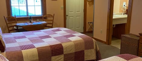 Ein Schlafzimmer mit zwei Betten, einem Schreibtisch und einem Stuhl. Es gibt ein Fenster mit Jalousien, eine Tür und ein Spiegel im Badezimmer.