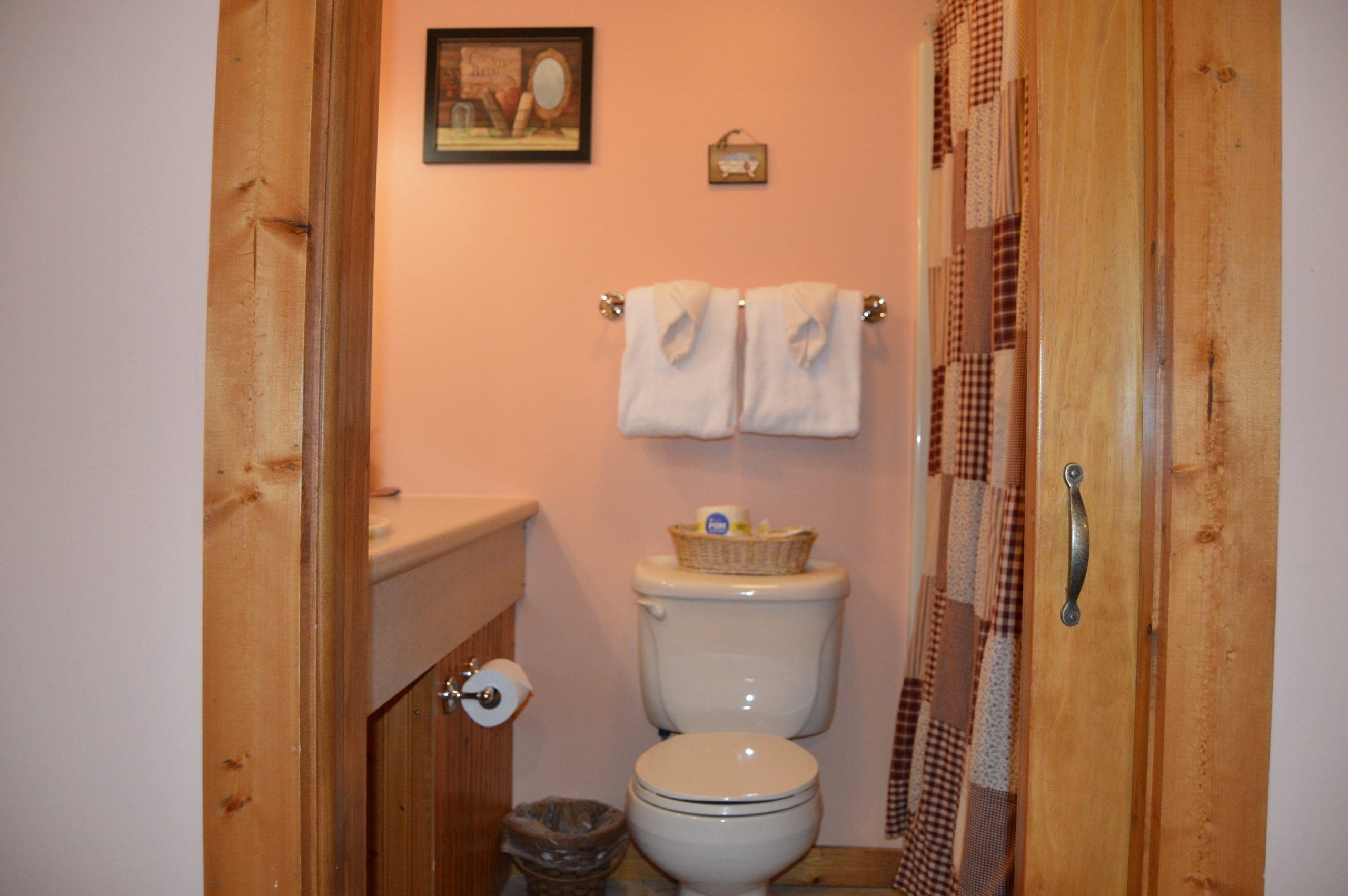 A small bathroom with a toilet, a roll of toilet paper, a basket on the toilet tank, and a towel rack with two towels.