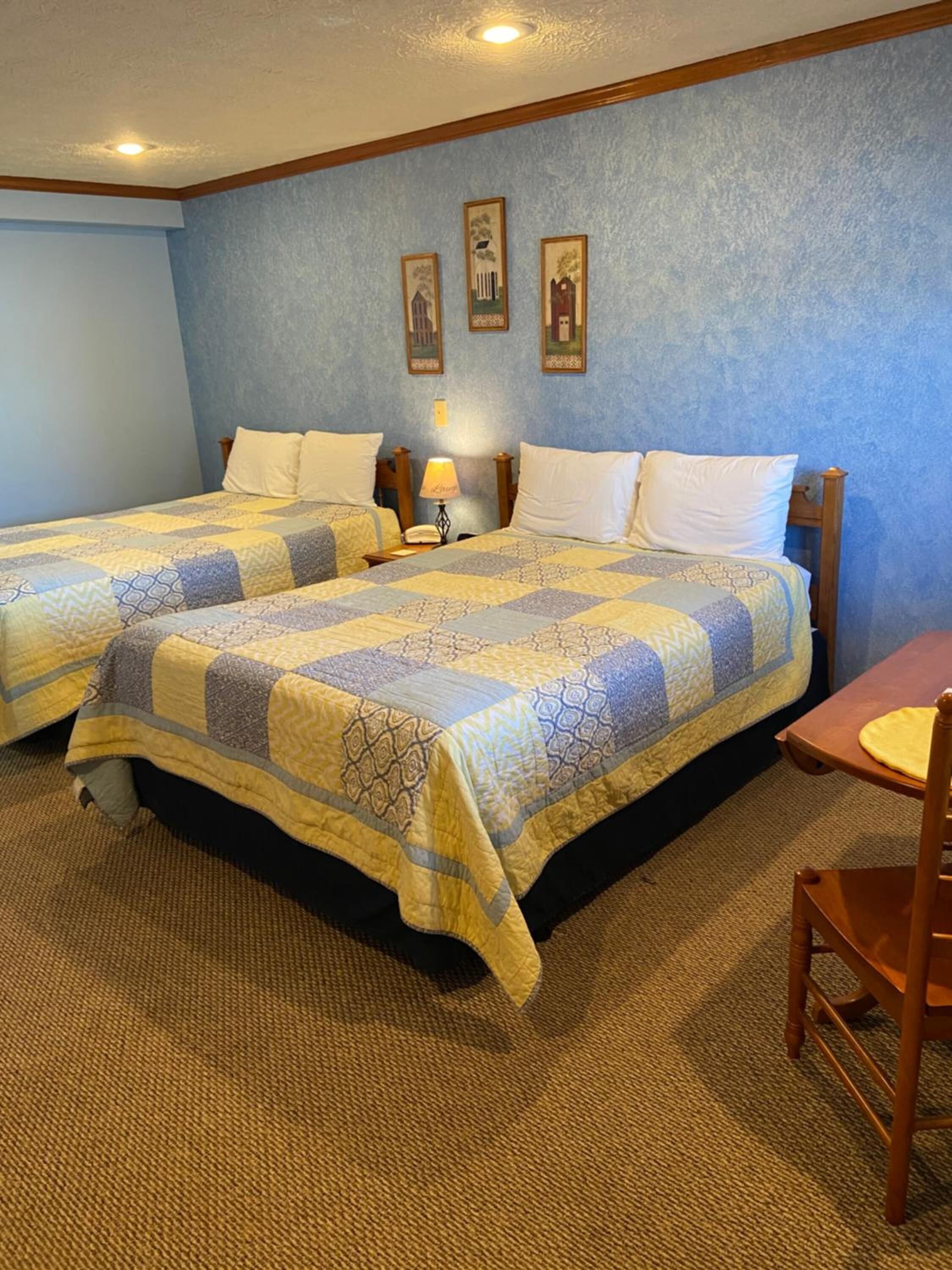 A hotel room with two beds, a small table, and a chair. There are three framed pictures on the wall.
