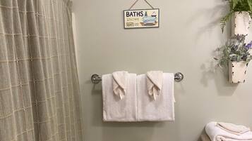 A bathroom with a toilet, a towel rack with two towels, a sign above the toilet, a basket on the toilet, a trash bin, and a plant on the wall.