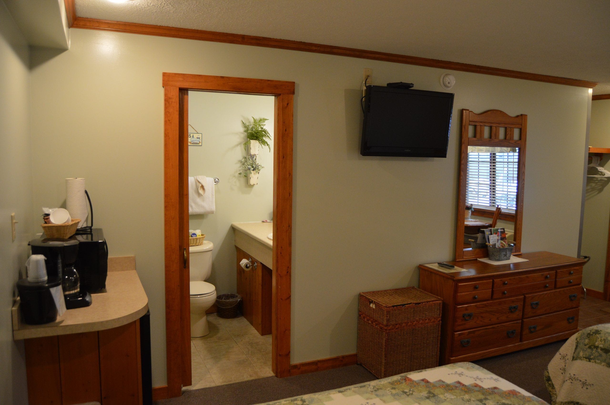 Une pièce de taille modeste, avec une commode en bois, un téléviseur fixé au mur, une petite fenêtre avec des stores et une salle de bain visible à travers une porte ouverte.