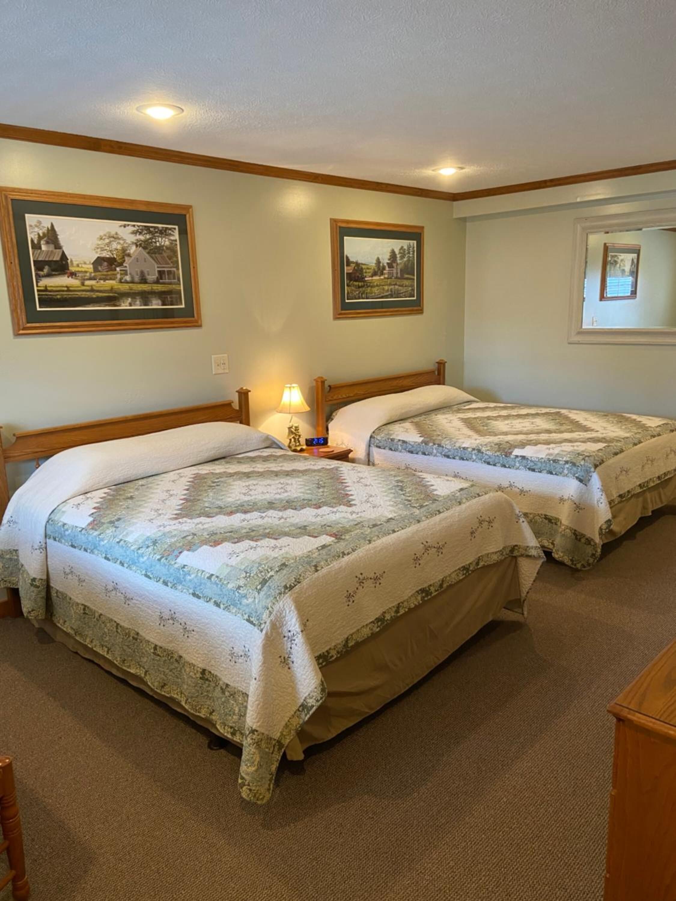 A room with two beds, each with a quilted bedspread, a nightstand with a lamp, and framed pictures on the walls.