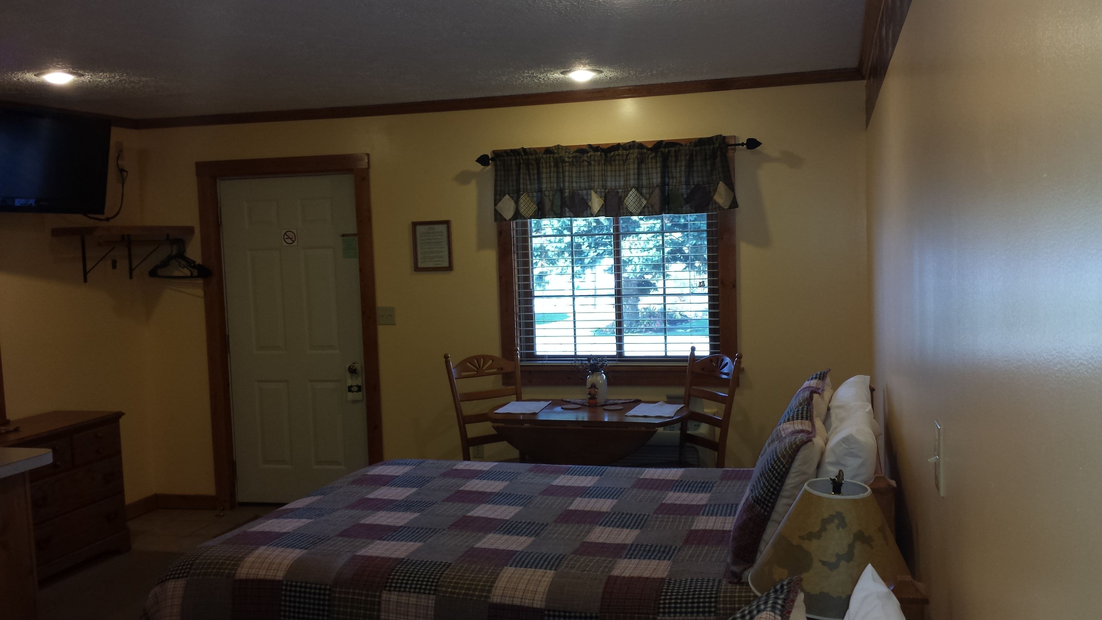 A bedroom with a bed, a table with chairs, a window with blinds, a door, and a television mounted on the wall.