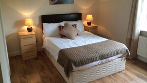 A neatly made bed with a headboard, two bedside tables with lamps, a painting of boats on a beach, and a window with curtains. - Doolin Village Lodges (Doolin)