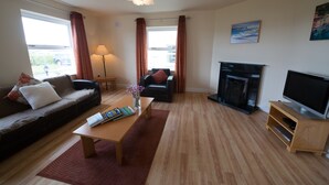 A living room with a sofa, coffee table, TV stand, fireplace, and large windows with curtains. - Doolin Village Lodges (Doolin)