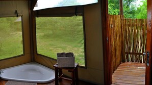 A room with a bathtub, a small table with a basket, and a view of a wooden deck and greenery outside. - Lion Tree Top Lodge (Hoedspruit)