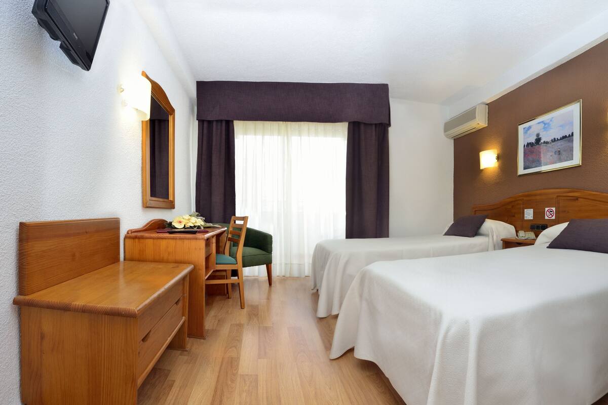 A hotel room with two beds, a desk, a chair, a TV, and a window with curtains.