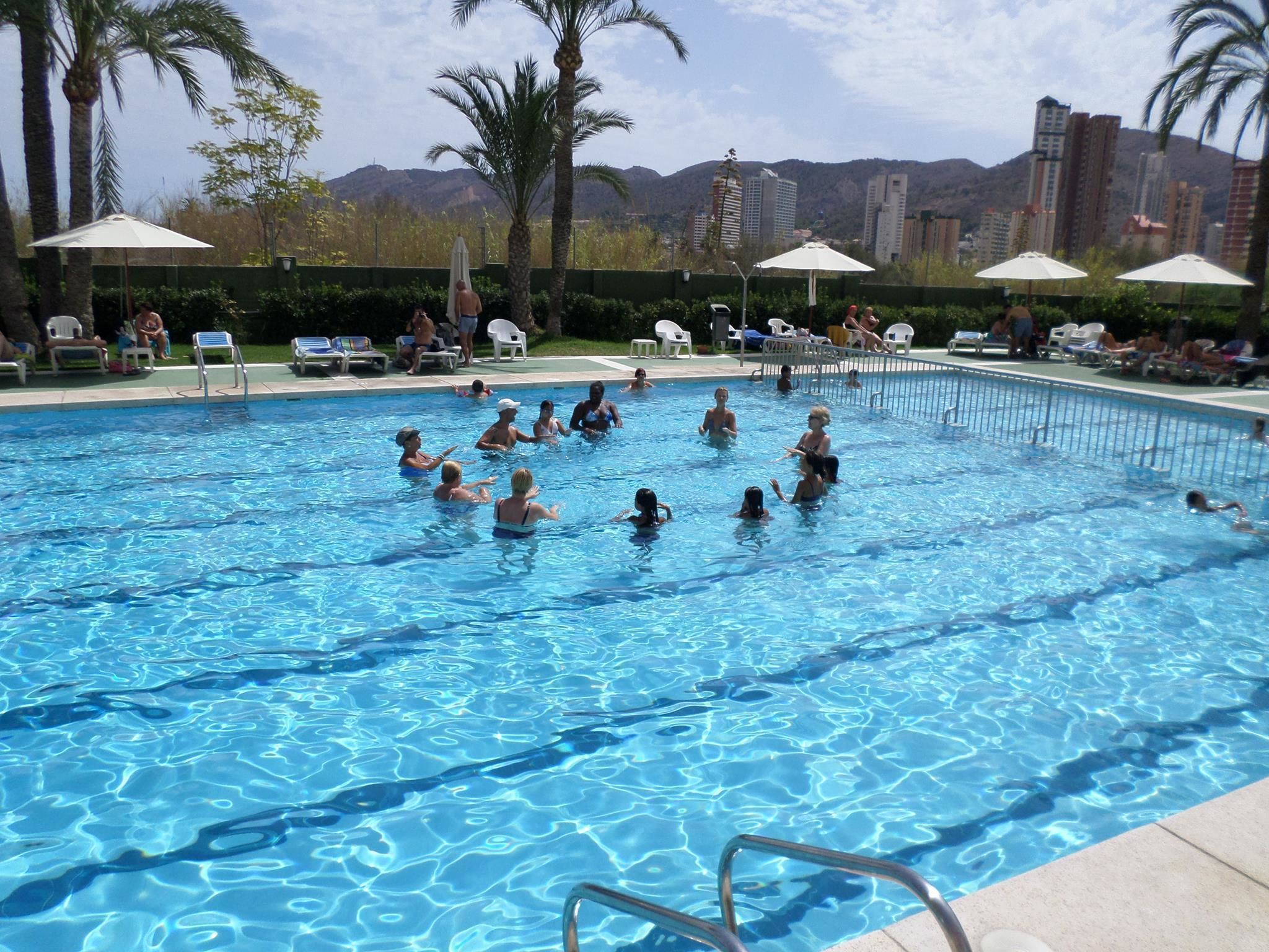 Ein Schwimmbecken mit darin badenden Personen, umgeben von Liegestühlen und Sonnenschirmen, mit einer Stadtkulisse im Hintergrund.