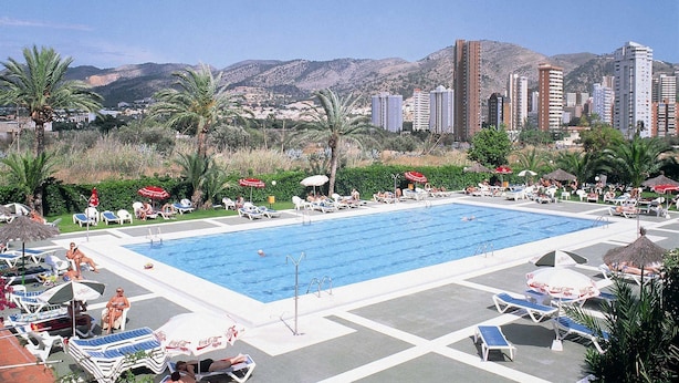 A pool area with lounge chairs and umbrellas, surrounded by palm trees and mountains in the background.