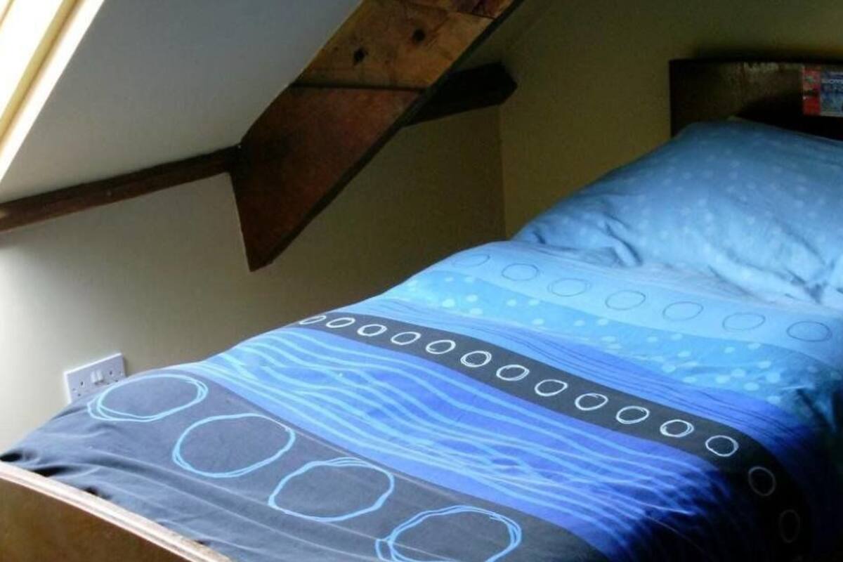 A single bed with a blue and white patterned bedspread, a wooden headboard, and a sloped ceiling with a window.